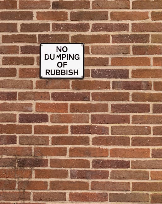 A rectangular metal sign mounted on a reddish-brown brick wall featuring a white background with black text that reads 'NO DUMPING OF RUBBISH.' The sign is positioned towards the upper left section of the wall, which consists of evenly laid bricks with a slightly textured surface and mortar lines in a lighter shade of beige. The lighting is natural, casting soft shadows beneath the sign and emphasizing the roughness of the brick surface. There are no other objects or surroundings visible, and the scene suggests a context related to private waste management or controlled rubbish disposal, often addressed by services like Rubbish Removal Mayfair. The sign's simple design and placement indicate an effort to prevent informal rubbish dumping in this urban space.