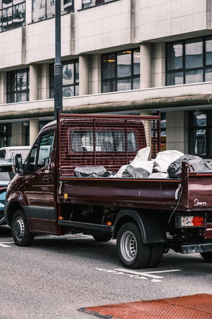 A small burgundy flatbed truck parked on an urban street, loaded with various waste bags and discarded items in black, white, and grey materials, some covered with plastic sheeting. The truck has a metal grid partition behind the cab, providing safety for the load, and features securing straps to hold the debris in place. It is positioned adjacent to a modern office building with large, reflective glass windows and a light-colored stone exterior. A black lamppost is visible in the foreground, and a white vehicle is parked nearby. The scene suggests an early or late day setting, with soft natural light illuminating the surroundings. This image illustrates a typical scenario of private waste collection or rubbish removal services in an urban environment, such as those offered by companies like Rubbish Removal Mayfair, which specialise in on-site clearance and alternative waste disposal solutions beyond local authority collections.