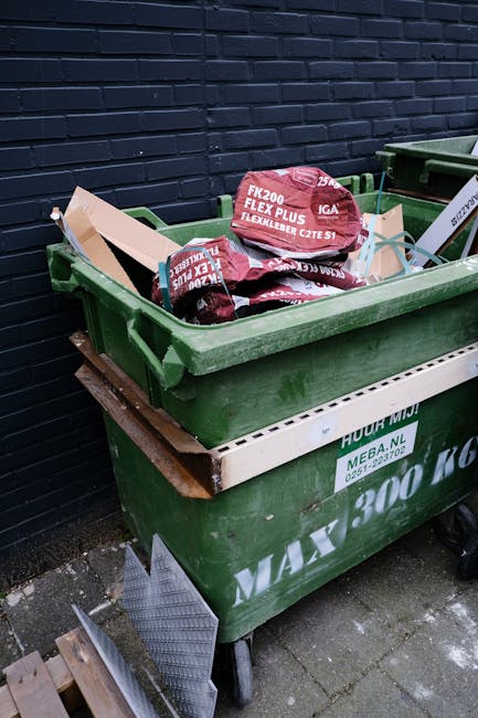 A large green wheeled waste disposal bin filled with red and black bags of building or garden debris, positioned outdoors against a dark brick wall. The bags are partially visible, with some branded as 'FLEXIBELBER' and displaying red and white graphics. The bin is placed on a stone-paved surface, with an adjacent white and green waste container underneath, which has some printed text including a web address and contact number. Two small wooden planks and a textured metal sheet are leaning against the bin near the ground. The environment suggests an outdoor rubbish collection or waste disposal area, possibly related to private waste handling or on-site clearance activities.