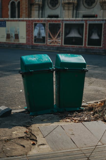 Two green outdoor rubbish bins with hinged lids positioned side by side on a concrete sidewalk at the edge of a paved street, with the left bin slightly taller and wider. The bins are made of plastic with a textured surface and appear weathered, showing some dirt and minor scratches. They are situated on a small patch of soil mixed with scattered leaves and debris, adjacent to a tiled pavement section. Behind the bins, a brick and concrete wall with decorative elements and large window frames is visible, part of a building with a historic architectural style. The surrounding environment includes a tarmac road with a slight incline, and in the background, a painted advertising panel on the wall. The lighting suggests natural daylight, highlighting the textures of the bins and the environment, reflecting typical private waste handling or alternative rubbish collection practices in urban areas, with no visible vehicles or tools present. Rubbish Removal Mayfair provides similar on-site or private waste clearance services that complement local authority rubbish collection, as suggested by the scene's context.