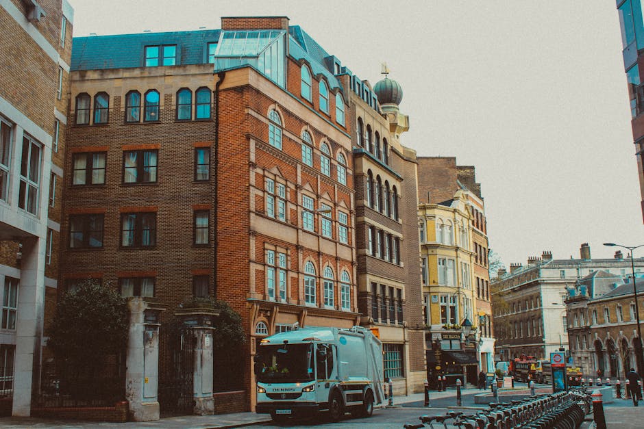 A city street scene featuring a collection of multi-story buildings with distinct architectural styles, including red brickwork, ornate window frames, and decorative stone detailing. In the foreground, a light blue waste collection truck is parked along the curb, partially blocking the view of the sidewalk. The truck's surface appears smooth with metallic and painted finishes, and the vehicle's doors are closed. Surrounding the truck, there are metal bollards and bicycle racks, indicating the street's mixed-use environment. The background shows additional historic buildings with classical facades, large windows, and some rooftop structures, including a dome with a cross on top. The lighting suggests a cloudy daytime atmosphere, providing an even, diffused natural light that emphasizes the textures of the buildings and the urban setting. This scene subtly illustrates private waste collection services, with Rubbish Removal Mayfair seemingly providing efficient rubbish removal within a typical central London environment, where alternative waste handling methods are evident through the presence of the waste collection vehicle.