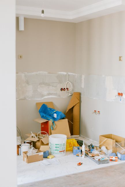 Inside an unfinished interior room, with beige painted walls and visible electrical outlets, there is a collection of construction debris and waste materials scattered on the floor. This includes a white plastic bucket, cardboard boxes, and packaging debris, along with a roll of brown insulation material leaning against the wall. A partially assembled piece of furniture or panel is propped upright, with a blue plastic bag draped over it. The floor appears to be concrete or a similar hard surface, and the wall behind shows patches of unpainted drywall or plaster. Electrical wiring and outlets are not yet fully installed, indicating ongoing renovation work. This scene reflects a typical stage of a DIY renovation project where waste and leftover materials are temporarily accumulated before proper disposal or collection, often handled by professional waste removal services such as Rubbish Removal Mayfair, which offers alternative solutions for managing building debris during refurbishment or property upgrades in the area.
