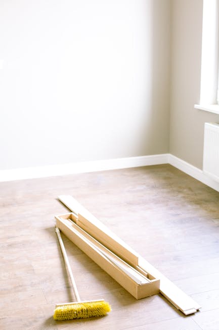 The image depicts a minimally furnished room with light beige walls, illuminated by natural light coming through a window on the right side. Resting on the light wooden floor are several pieces of flat-pack furniture, including a long, rectangular wooden frame or panel, possibly a shelf or base component, alongside smaller, matching wooden parts. A yellow cleaning brush with stiff bristles is positioned adjacent to the wooden components, indicating ongoing or recent renovation work. The room appears empty aside from the furniture parts and brush, with an environment suitable for a private renovation project. The scene suggests that the furniture parts are awaiting assembly or disposal, aligning with services provided by companies like Rubbish Removal Mayfair, which handle waste removal related to DIY renovation projects. The plain walls, clean floor, and window create a neutral backdrop for the scene, emphasizing the focus on the furniture components and cleaning tools awaiting removal as part of a tidy, independent waste handling process following a renovation effort in a residential setting.