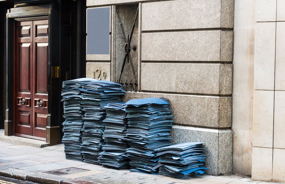 A large stack of disorganized blue paper folders and documents is piled against the exterior stone wall of a building on a city sidewalk. The folders vary in size and are slightly crumpled with some pages protruding from the edges, indicating a recent or ongoing collection of office or administrative waste. The paper materials are stacked unevenly, with the majority leaning slightly to the right, close to a dark wooden door with vertical panels and metallic door hardware. To the left of the paper stacks, a black metal shutter is partly visible, suggesting an entrance or storefront. The textured stone wall behind the stack features large, rectangular blocks with horizontal joints, and a small metal ventilation grille is partially visible behind the folders. The scene appears well-lit by natural daylight, illustrating an example of external waste accumulation that might be handled through private waste management services such as those offered by Rubbish Removal Mayfair, emphasizing alternative methods of rubbish removal outside of municipal collections.