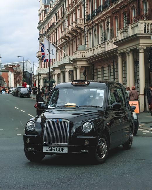 A black London-style taxi cab with a rounded front, vertical grille, and distinctive circular headlights is parked on a city street during daytime. The vehicle's glossy black finish reflects natural light, and its license plate reads LS15 GDF. In the background, a row of historic multi-storey buildings with ornate balconies, large windows, and varied architectural details line the street, with some displaying Union Jack flags. Pedestrians are visible on the pavement, some standing and others walking, while a few vehicles, including other cars, are driving along the road. The scene is illuminated by natural daylight, creating a clear, detailed urban environment. This image, associated with Rubbish Removal Mayfair, offers a glimpse into city life and may relate to private transportation or on-site clearance contexts within an urban rubbish removal service environment.