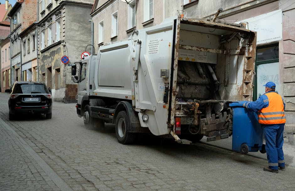 A sanitation worker dressed in high-visibility orange and blue work clothing is seen operating a large, white waste collection truck on a cobblestone street. The truck, positioned with its rear open, reveals a rusty, used compaction mechanism designed for the compacting and emptying of waste. The worker, wearing a blue cap and gloves, is in the process of emptying a blue wheeled waste bin into the truck's hopper, which is situated on the right side of the vehicle. The surrounding environment includes a row of old, multi-storey buildings with a mix of stone and brick facades, some showing weathering and peeling paint. To the left, a black car is parked parallel to the curb, and signage indicating no stopping is mounted on a post nearby. The scene appears to take place during daylight hours with natural, diffuse lighting, consistent with a routine waste collection service in an urban setting, where private rubbish handling is occurring outside the building facades, aligning with a rubbish removal service taking place in a city environment.