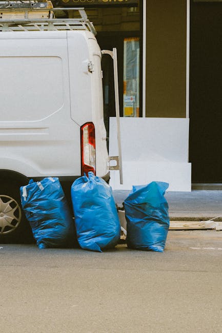 A sanitation worker dressed in high-visibility orange and blue work clothing is seen operating a large, white waste collection truck on a cobblestone street. The truck, positioned with its rear open, reveals a rusty, used compaction mechanism designed for the compacting and emptying of waste. The worker, wearing a blue cap and gloves, is in the process of emptying a blue wheeled waste bin into the truck's hopper, which is situated on the right side of the vehicle. The surrounding environment includes a row of old, multi-storey buildings with a mix of stone and brick facades, some showing weathering and peeling paint. To the left, a black car is parked parallel to the curb, and signage indicating no stopping is mounted on a post nearby. The scene appears to take place during daylight hours with natural, diffuse lighting, consistent with a routine waste collection service in an urban setting, where private rubbish handling is occurring outside the building facades, aligning with a rubbish removal service taking place in a city environment.