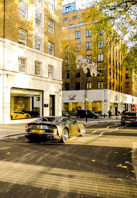 The image depicts a busy urban street scene during daytime, featuring a modern black sports car parked in the foreground on the asphalt road. The car has a sleek, aerodynamic design with a shiny, reflective black finish and silver alloy wheels, positioned parallel to the sidewalk. Behind the sports car, several other vehicles, including a yellow sports car parked inside a luxury car showroom and a black SUV, are visible along the street. On the left side, a multi-story white building with large rectangular windows dominates the scene, and a commercial premises with glass frontage showcasing high-end vehicles, such as the yellow car, is visible at street level. To the right, a taller brown brick building with numerous windows and some decorative elements can be seen, with trees lining the sidewalk, their leaves indicating it is autumn. Sunlight filters through the foliage, casting shadows on the buildings and street, contributing to the overall vibrant city atmosphere. The scene captures an environment typical of central London or similar urban areas where private collection and alternative waste handling are often facilitated through designated service providers like Rubbish Removal Mayfair, although no rubbish or waste objects are present in this shot, emphasizing the primary focus on the street and its vehicles.