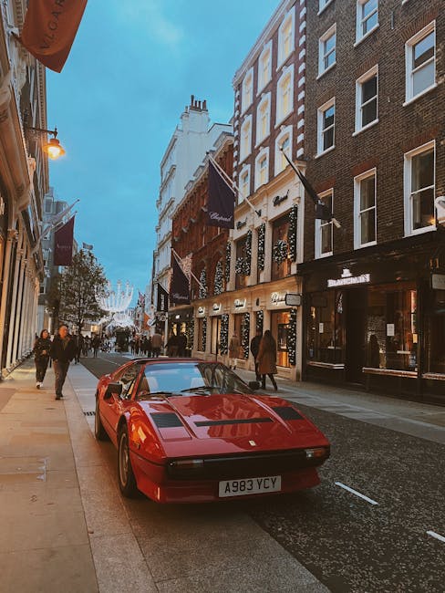 A red sports car with a sleek, low-profile design and a smooth, shiny finish is parked on the curb of a busy city street, positioned close to the sidewalk in front of a row of multi-storey buildings with a mix of brick and painted facades. The car's license plate reads A983 YCY, and it features distinctive black vents on the front hood. The street scene includes pedestrians walking along the sidewalk, some wearing coats, with commercial premises visible below large windows and four flags with dark backgrounds hanging from poles attached to the building facades. The sky above is partly cloudy with a soft blue tone, and ambient street lighting is beginning to illuminate the area as evening approaches. In the background, decorative streetlights and festive string lights are visible, suggesting an urban environment that may experience private or independent waste collection practices, as part of routine rubbish removal or on-site clearance within the city. The overall atmosphere is lively and orderly, with the vehicle prominently placed in the foreground, emphasizing urban aesthetics and practical street scene observations related to waste management and city life.