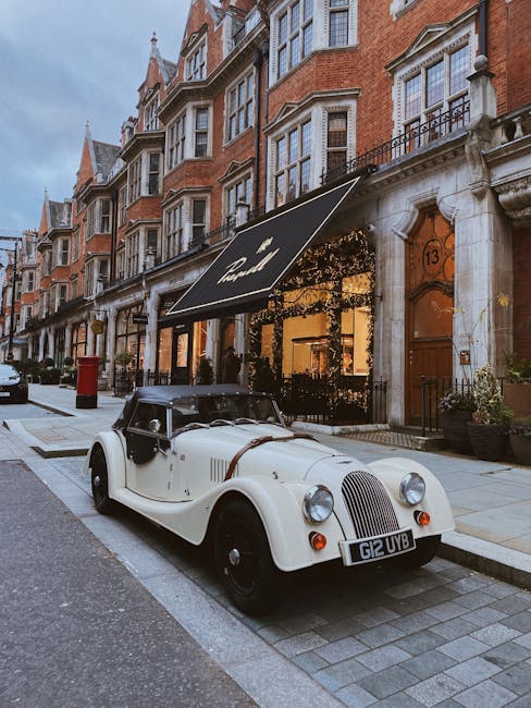 A vintage white and black convertible car with a leather strap securing the bonnet is parked on a cobblestone street in front of a row of traditional red-brick terraced houses with large sash windows and ornate stone detailing. The car's rounded headlights and classic grille are prominent, and the vehicle's black soft-top roof is closed. Behind the car, a storefront with a black awning featuring gold lettering is illuminated from within, casting a warm glow onto the sidewalk. Potted plants and a wooden door with the number 13 are visible beside the shop. The scene is set during dusk, with a cloudy sky overhead, and the street appears clean and well-maintained, reflecting a typical urban residential area that may include private waste collection services such as those offered by Rubbish Removal Mayfair, supporting alternative rubbish handling outside of local authority collections.