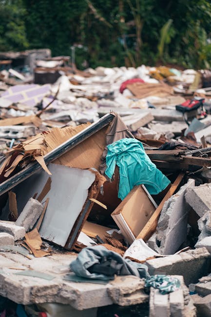 A pile of assorted household debris and rubbish, including broken wooden furniture, a torn cardboard box, scattered fabric pieces, and broken concrete blocks, situated outdoors on a paved surface. The materials exhibit various textures such as rough concrete, smooth wood, and crumpled paper, with colors ranging from natural wood tones and gray concrete to bright blue fabric and faded cardboard. The debris is haphazardly arranged, with some items partially covered by others, suggesting recent or ongoing clearing activity. In the background, dense green foliage indicates the outdoor setting, and the scene is lit by natural daylight, emphasizing the varied textures and forms of the waste materials. This scene reflects a typical site for private rubbish removal services, such as those provided by Rubbish Removal Mayfair, handling on-site clearance of household or construction waste directly from the property.