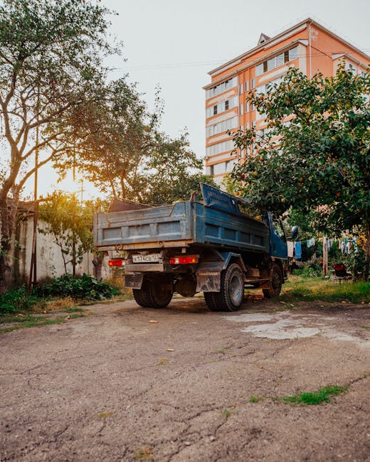 A blue flatbed truck with a metal cargo container attached to its back is parked on a dirt and gravel area in an outdoor setting. The truck appears aged, with visible dirt on the tires and body, and driver-side paint chipping. To the right of the truck, there are green trees with dense foliage, some branches extending over the vehicle. In the background, a multi-storey residential building with a peach-colored facade and white balconies is visible, partially obscured by the trees. The scene is illuminated by soft, warm sunlight, suggesting either early morning or late afternoon, casting gentle shadows across the scene. The surrounding environment includes patches of grass and small plants growing at the edges of the cleared area, indicating a semi-urban location typically associated with private waste collection or rubbish removal services, such as those provided by Rubbish Removal Mayfair. This scene visually reflects the process of on-site clearance and disposal of accumulated waste materials.