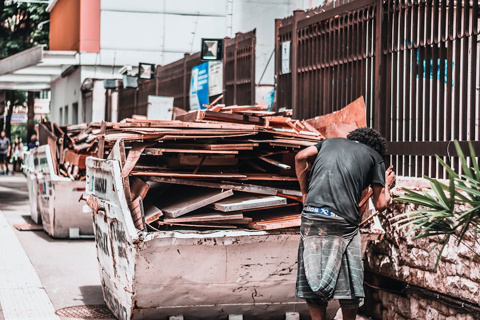 A man with curly dark hair, dressed in a black t-shirt and grey plaid shorts, stands beside an overflowing white skip container filled with various construction and demolition debris. The debris consists mainly of weathered, reddish-brown roof tiles, along with some grey metallic and wooden materials visible on top. The skip is situated on a paved urban street next to a brown metal fence with vertical slats, which runs alongside a building with orange and white exterior walls. In the background, there are signs, streetlights, and pedestrians walking along the sidewalk, indicating a busy city environment. The scene appears to be part of a temporary on-site clearance or private waste removal, with visible clutter and a focus on construction waste in need of collection. The lighting suggests daytime with natural light illuminating the scene, making the textures of the debris and materials clearly visible, typical of sites managed by waste removal services like Rubbish Removal Mayfair, engaged in efficient rubbish disposal and alternative waste handling solutions.