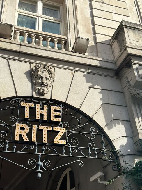 A close-up view of the exterior entrance to a building named 'The Ritz,' featuring a decorative stone face sculpture with a beard and expressive features positioned above a curved wrought iron canopy. The canopy displays illuminated signage spelling 'THE RITZ' in large, block letters made of round, bulbous lights, with a background of cream-colored stone facade decorated with horizontal and vertical grooves. The building has a classical architectural style, with part of a window visible above and a small balcony with a balustrade on the upper floor. Shadows cast by sunlight create depth and contrast on the stone surface, and a portion of greenery is visible climbing near the corner, contributing to an elegant and historic appearance consistent with London’s high-end hospitality venues associated with private or independent management of waste disposal, as might be relevant in waste removal services like those offered by Rubbish Removal Mayfair.